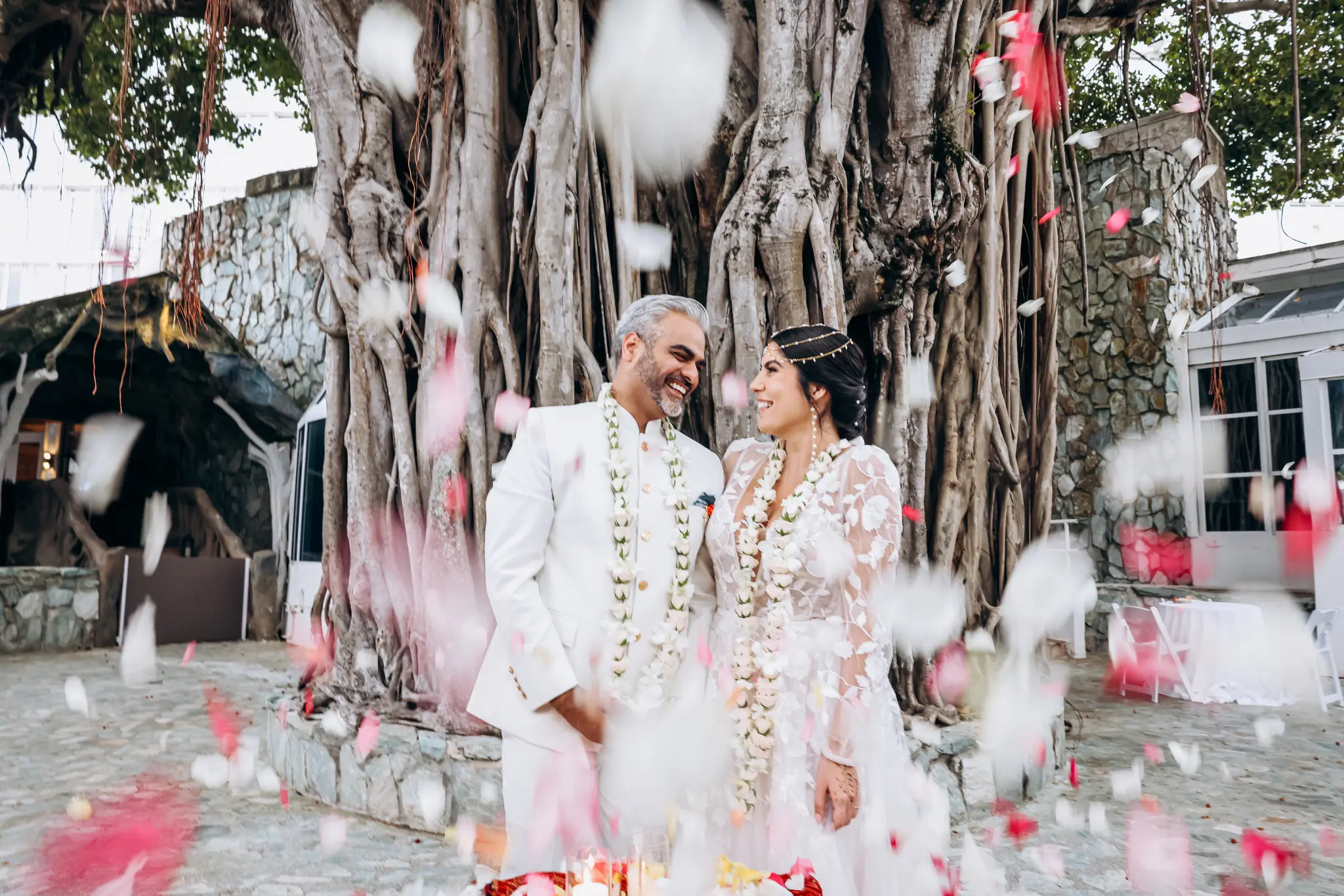 Newly married couple in formal attire wearing floral garlands as white and pink petals fall