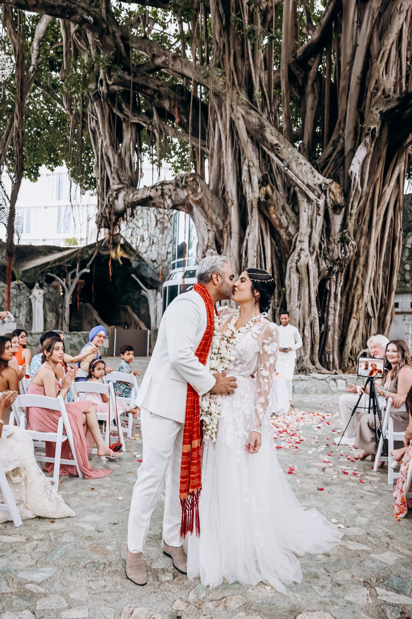 Couple kissing at an outdoor ceremony beneath a large banyan tree with guests along a petal-covered aisle
