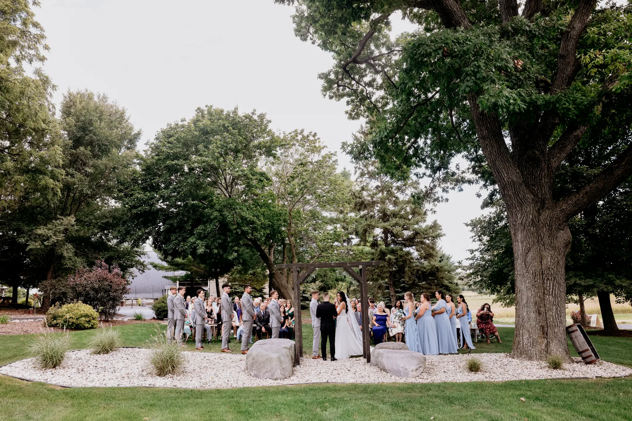 Wide garden wedding ceremony beneath a wood arbor with a full wedding party and guests