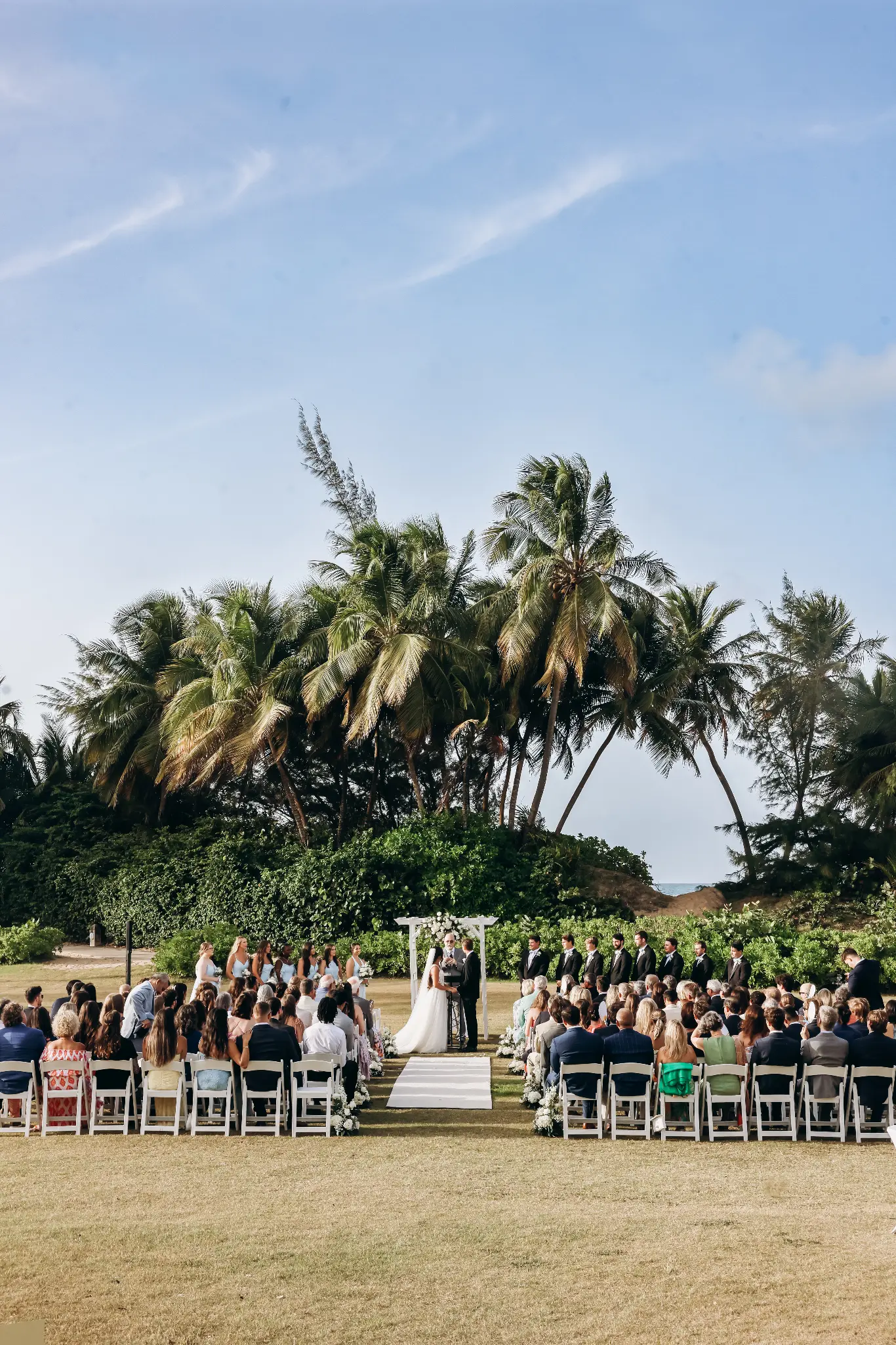 Coastal lawn wedding ceremony with a white arbor, formal wedding party, seated guests, and palm trees