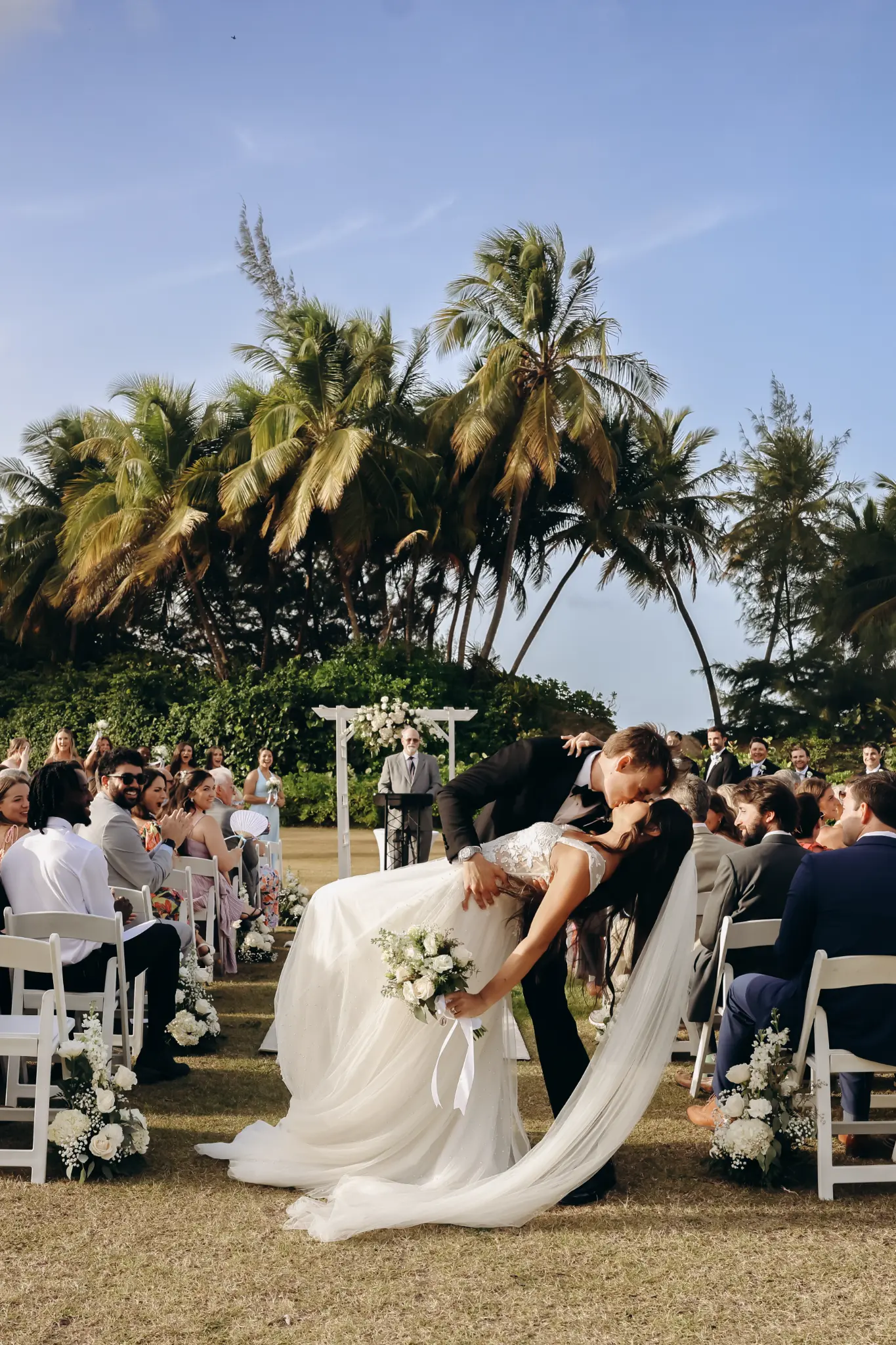 Wedding dip kiss during a palm-lined outdoor ceremony with guests along the aisle