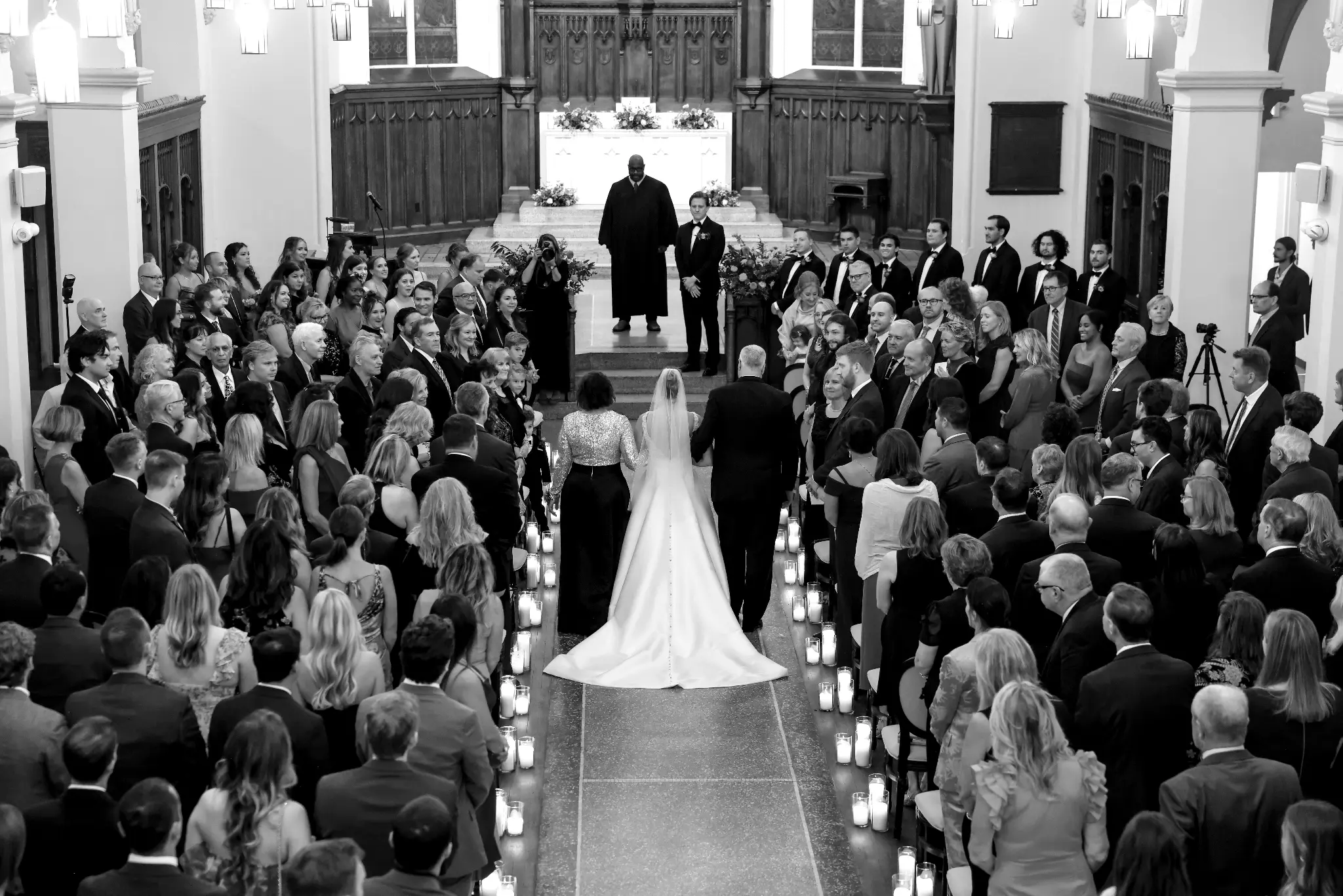 Black-and-white overhead view of a bride entering a candlelit church aisle with two escorts