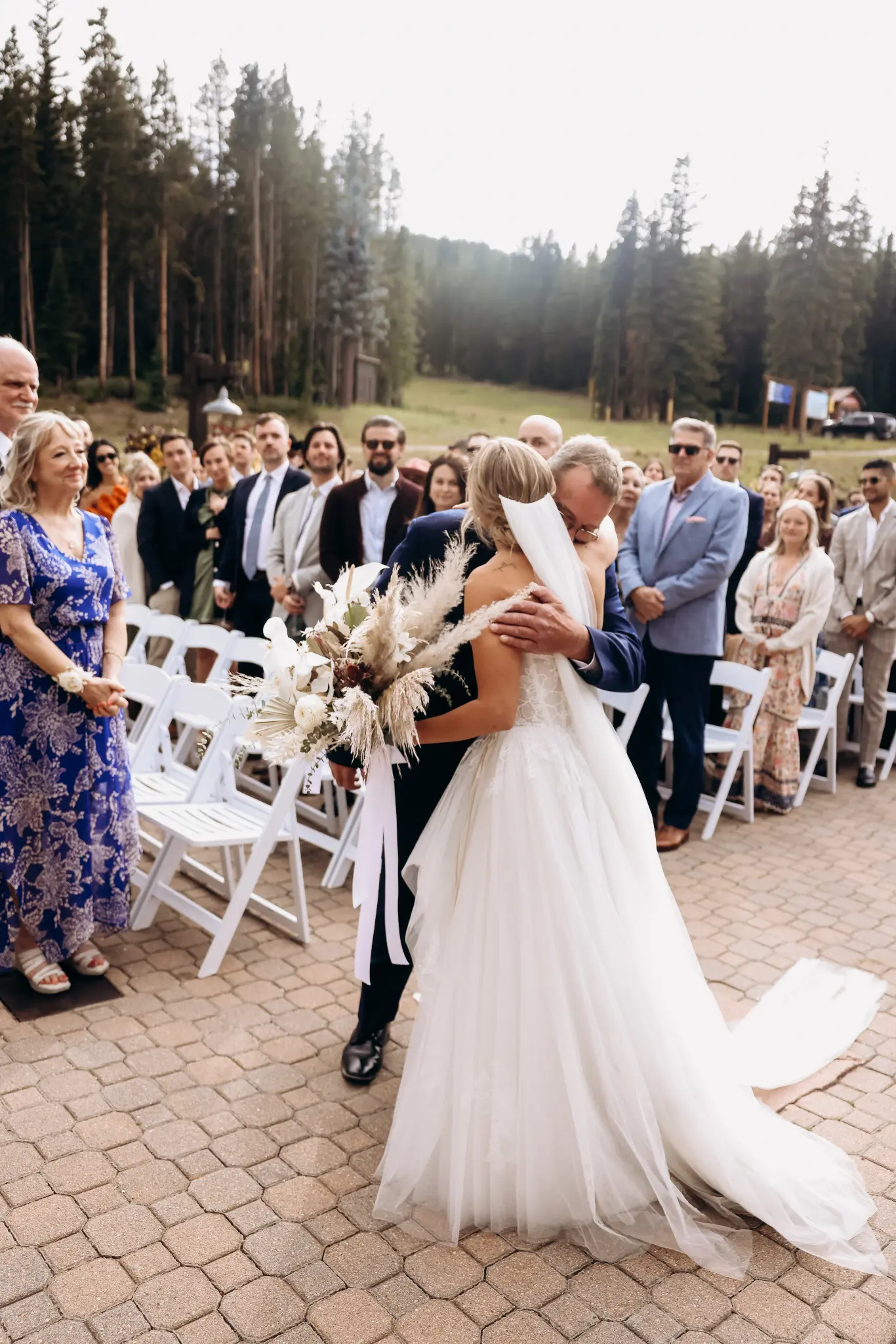 Bride and escort embracing at an outdoor ceremony with guests behind them