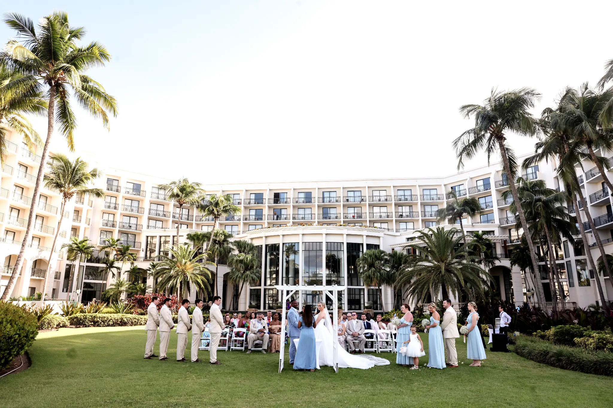 Outdoor palm-lined lawn wedding ceremony with a white arbor and wedding party in pale blue attire