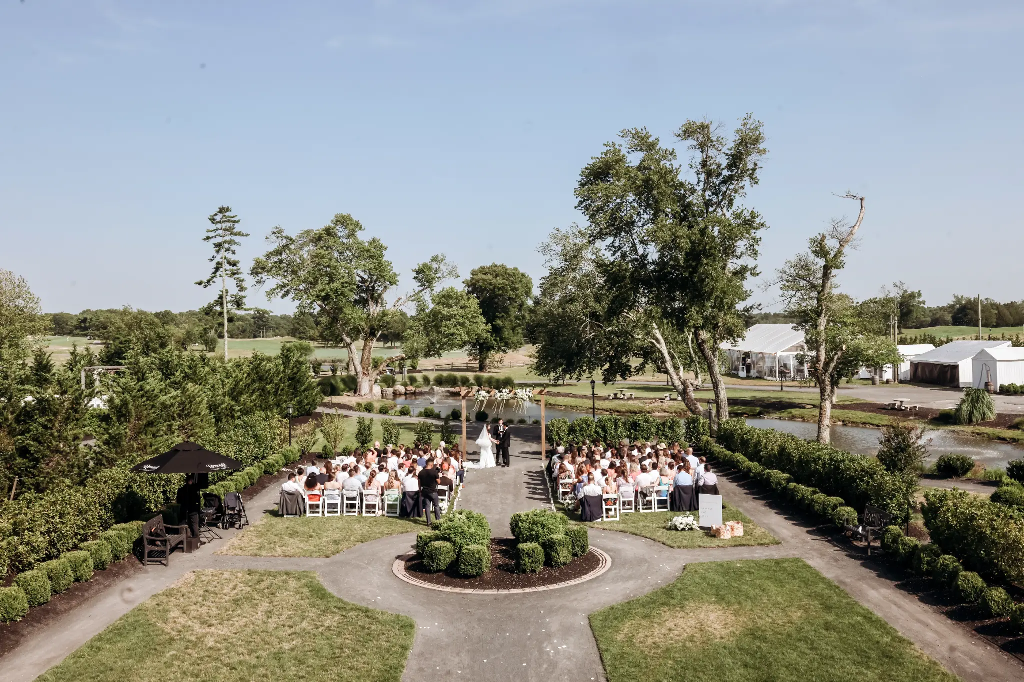 Elevated view of a garden wedding ceremony beneath a wood arbor beside a pond