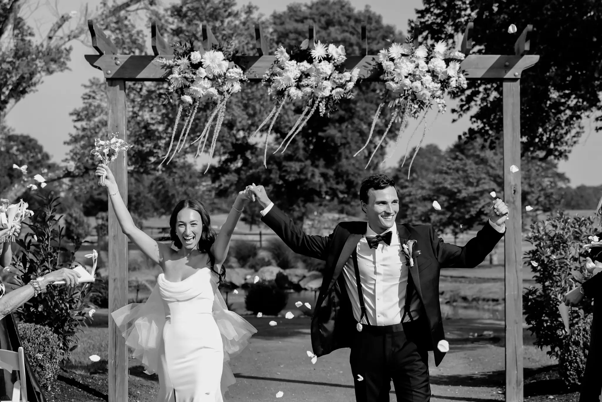 Black-and-white couple recessional beneath a flower-covered timber ceremony structure as guests toss petals