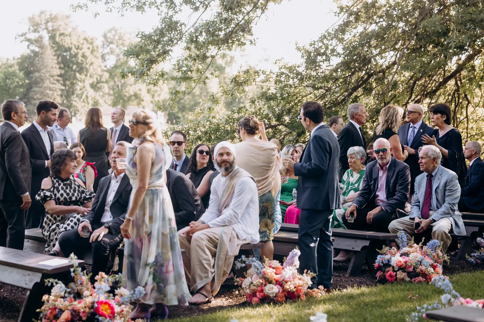 Guests gathering beneath trees before an outdoor ceremony with colorful aisle flowers