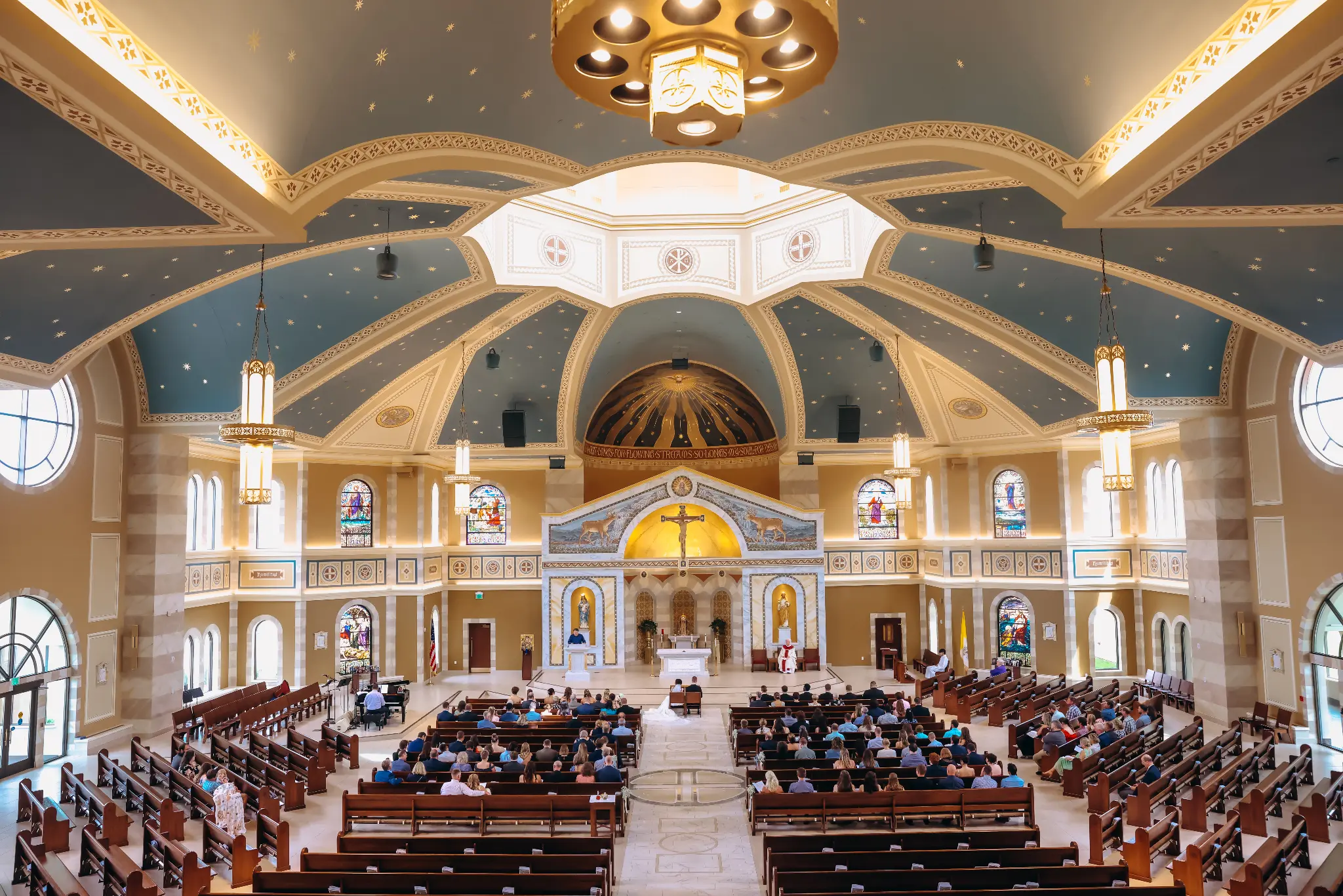 Wide view of a church ceremony with stained glass, a blue star-patterned ceiling, central altar, and guests in pews