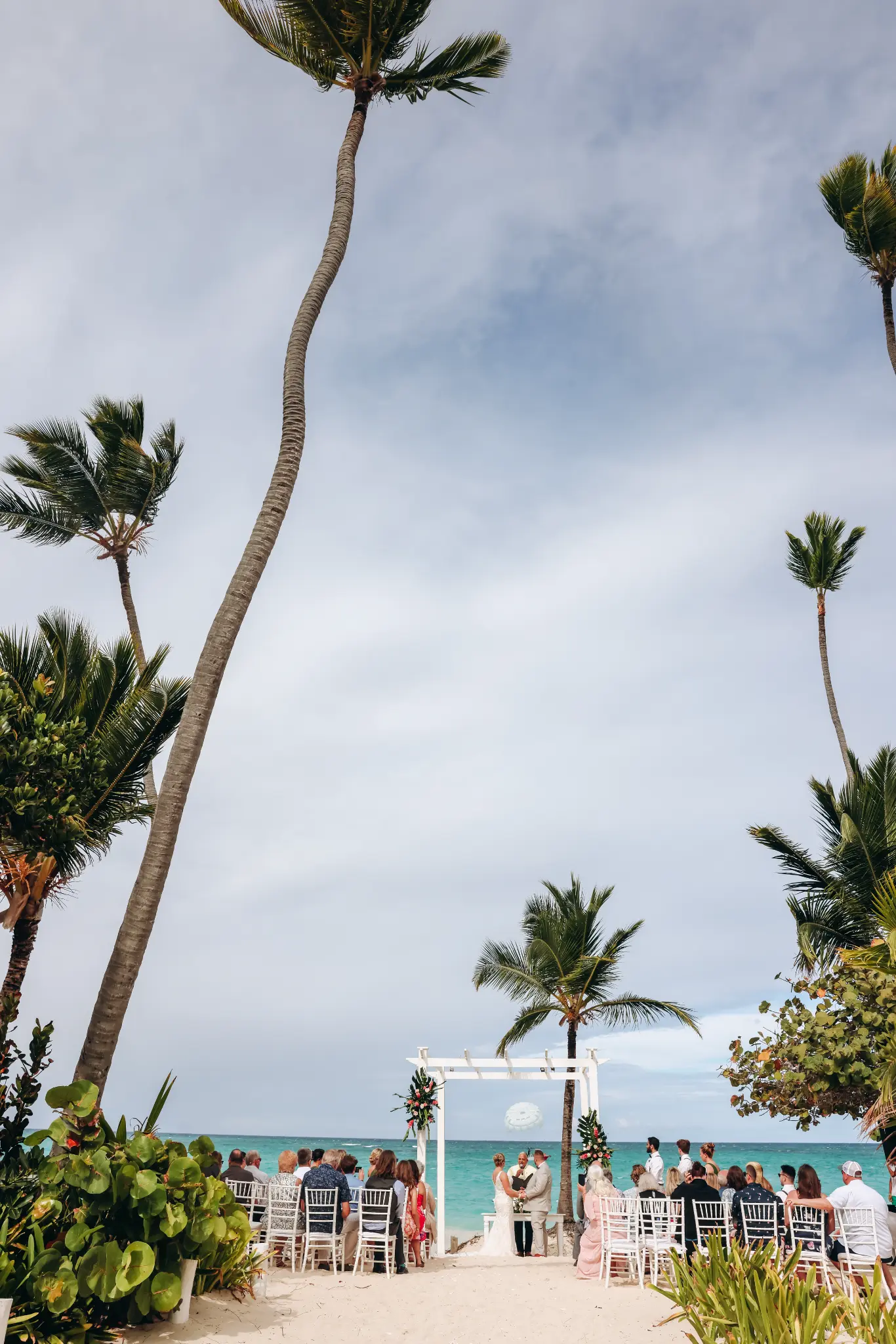 Beach wedding ceremony facing turquoise water, framed by palms and a white arbor