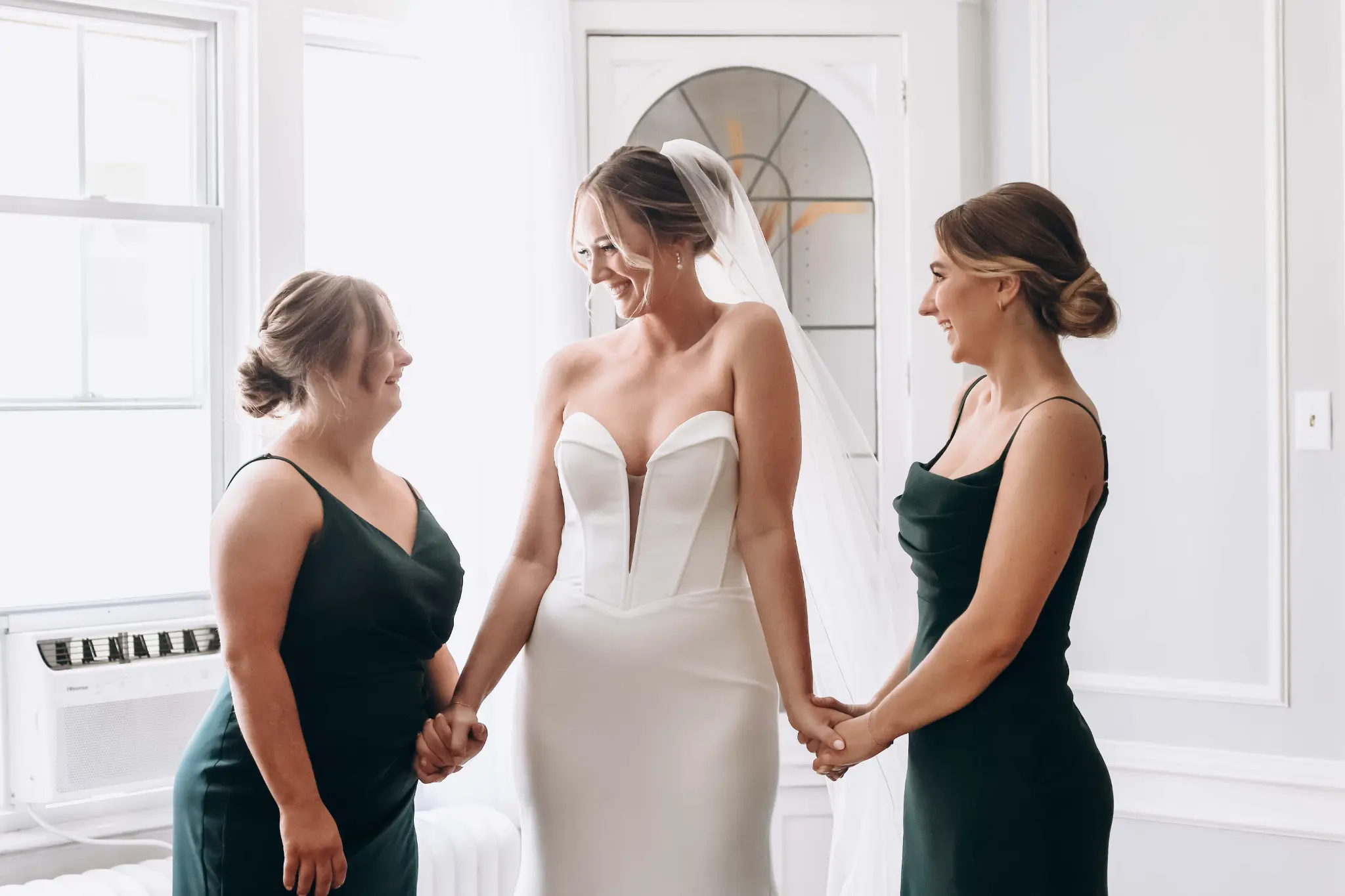 Veiled bride holding hands with two attendants in a bright room during wedding preparations