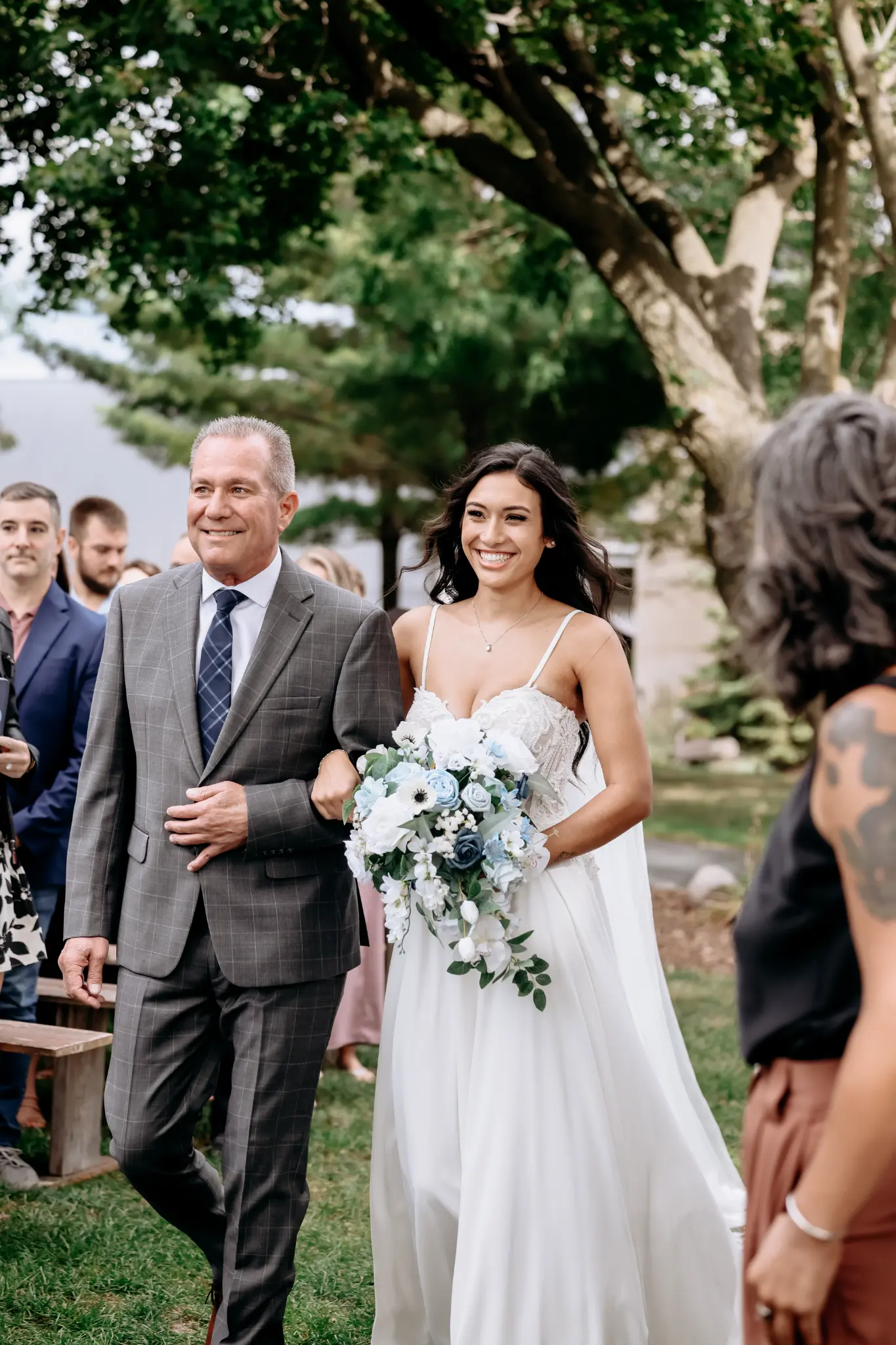 Bride walking down an outdoor wedding aisle with an escort and blue-and-white bouquet