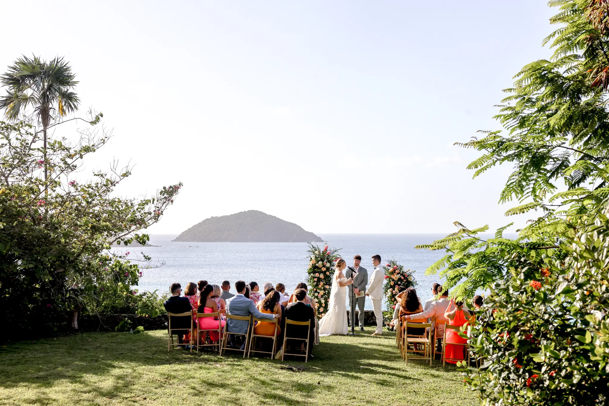 Coastal wedding ceremony overlooking the water and a small island, with guests seated below floral arrangements