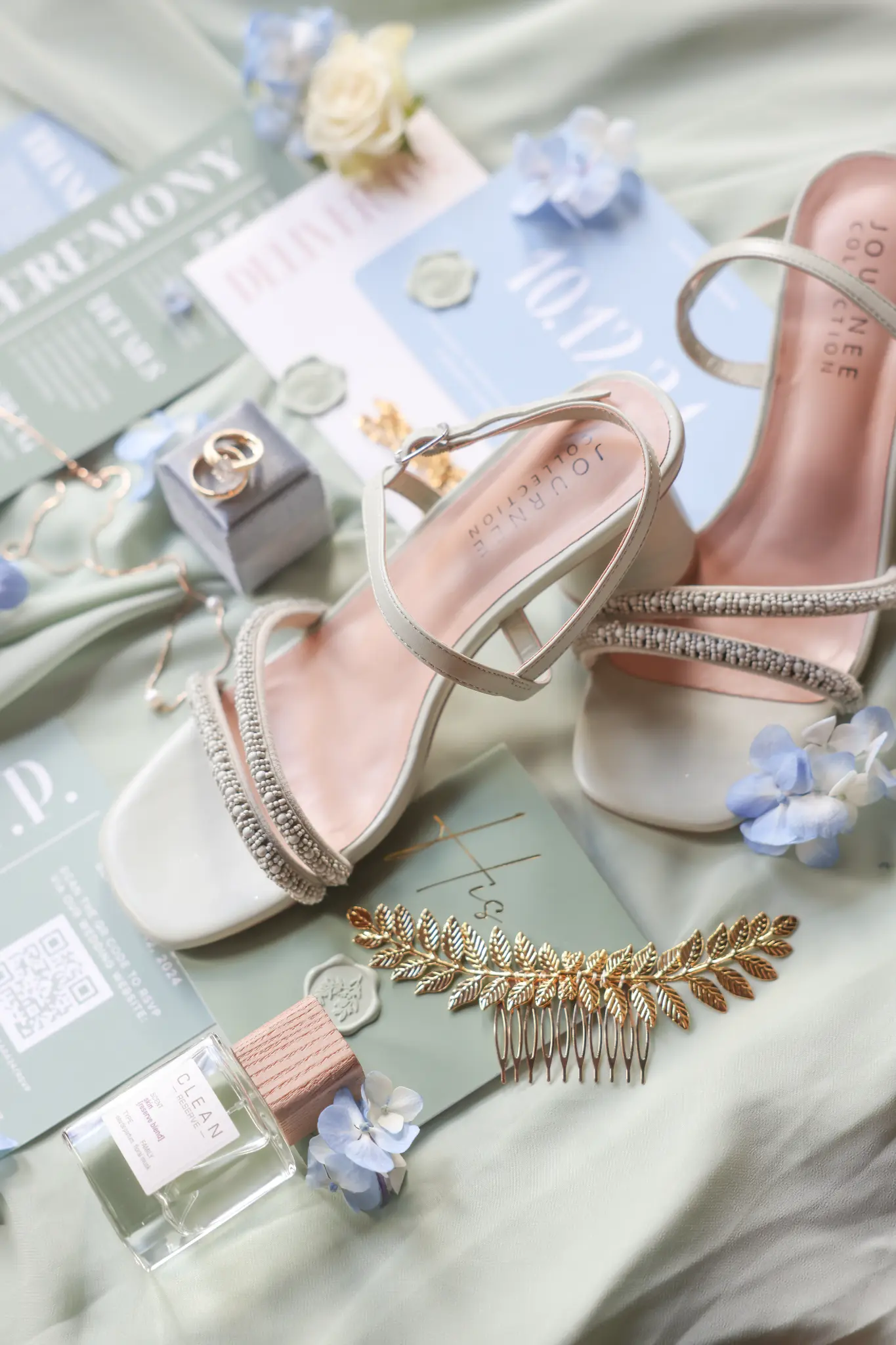 Bride’s embellished shoes, rings, hairpiece, stationery, and fragrance arranged on pale blue fabric