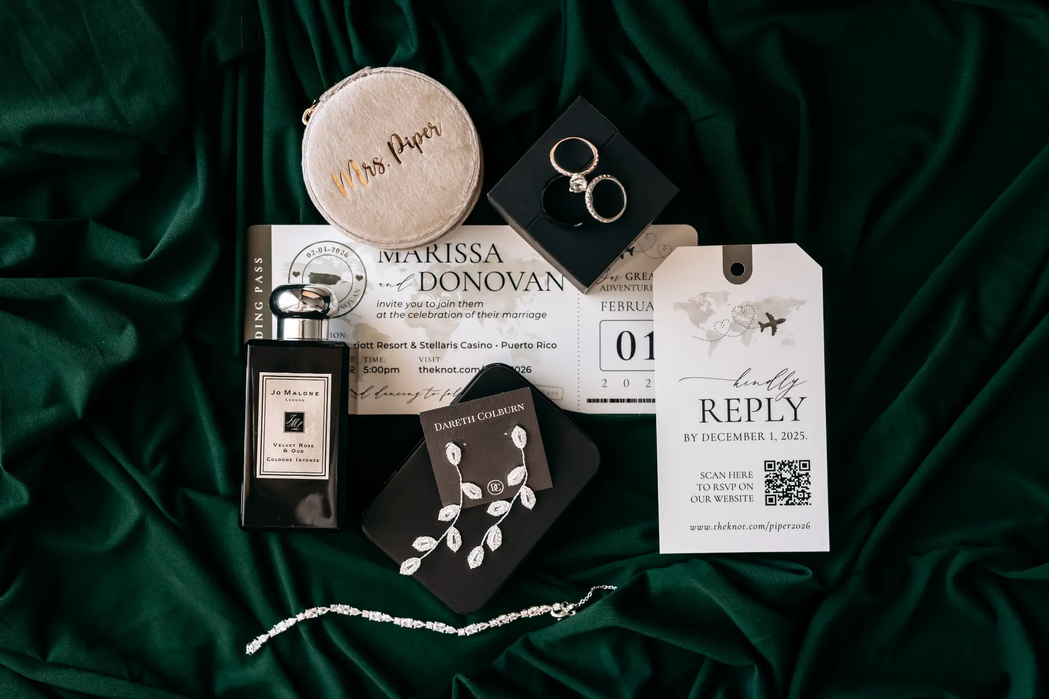 Bride’s wedding-day details with rings, invitation, fragrance, earrings, and reply card arranged on emerald fabric