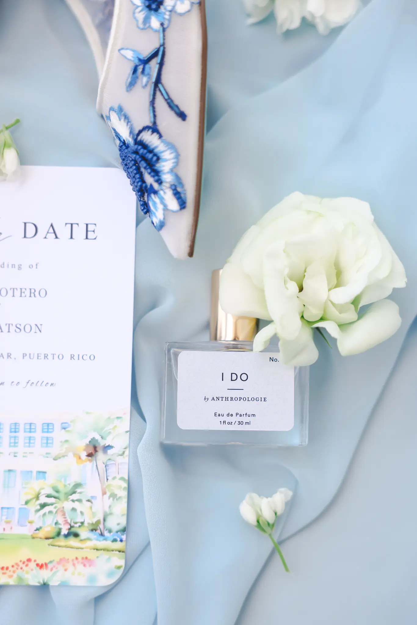 Bride’s embroidered shoe, invitation, white flower, and fragrance arranged on pale blue fabric