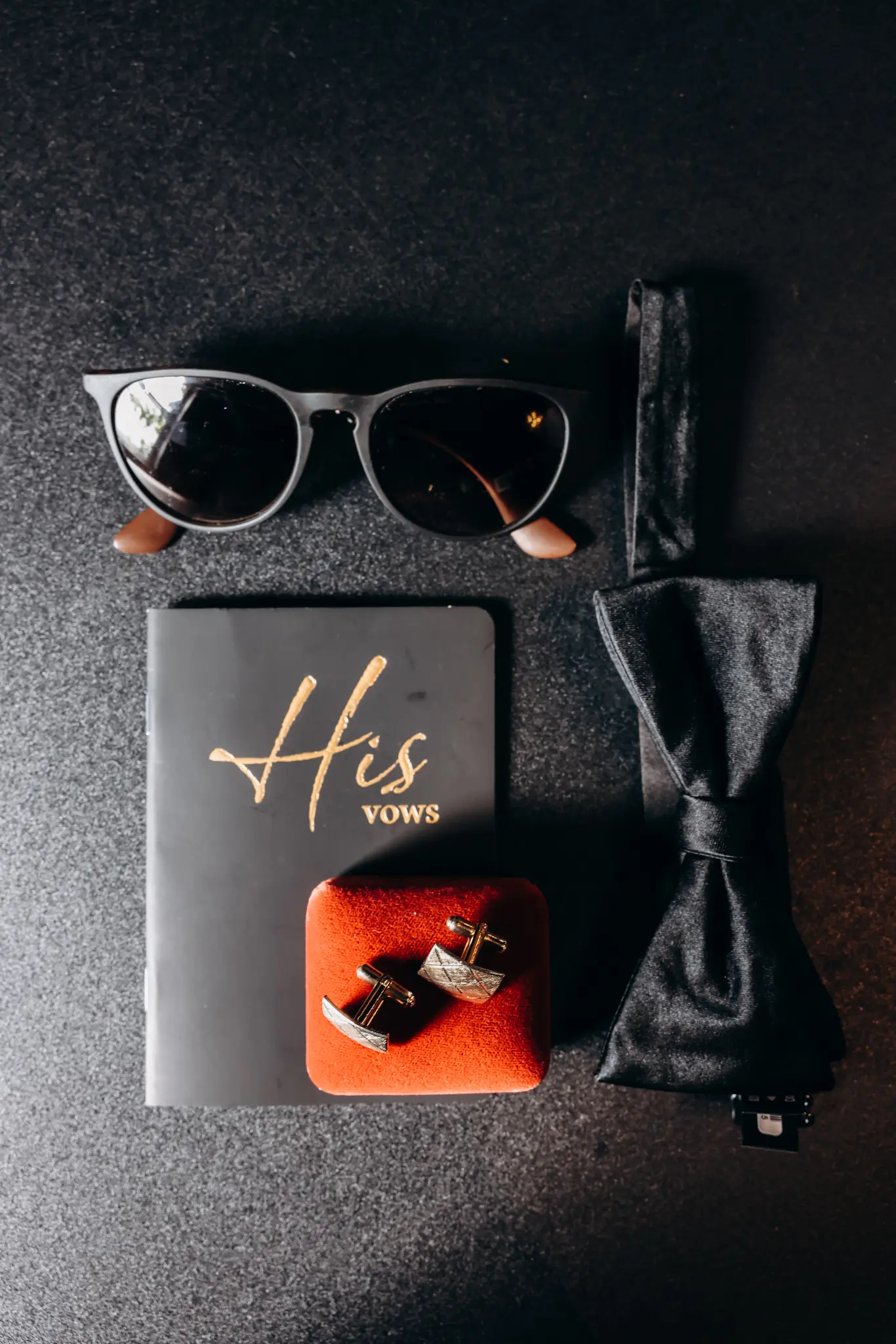 Groom’s wedding-day details with sunglasses, cufflinks, bow tie, and vows booklet