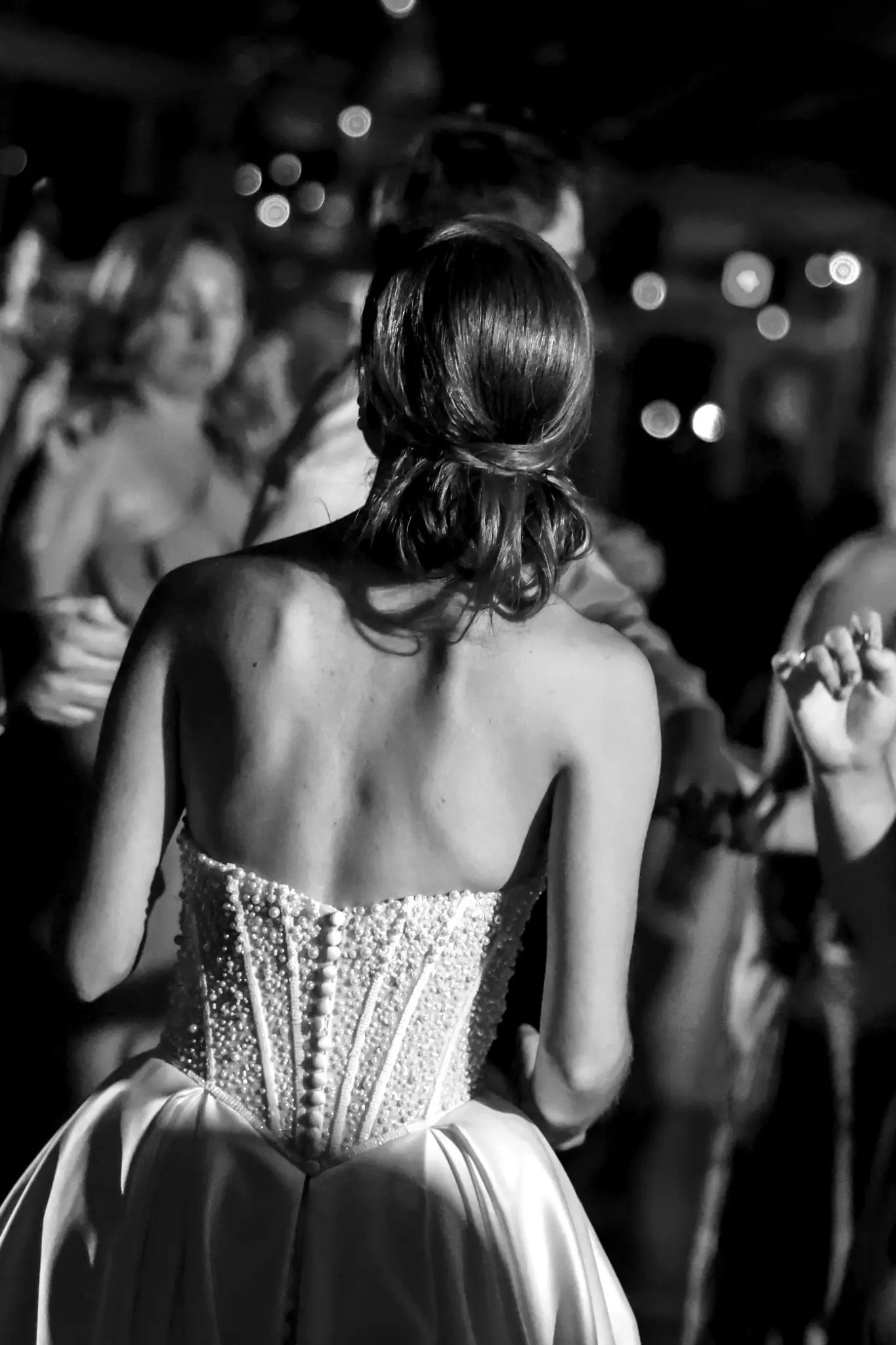 Black-and-white back view of a bride in a corseted gown moving through a wedding reception crowd