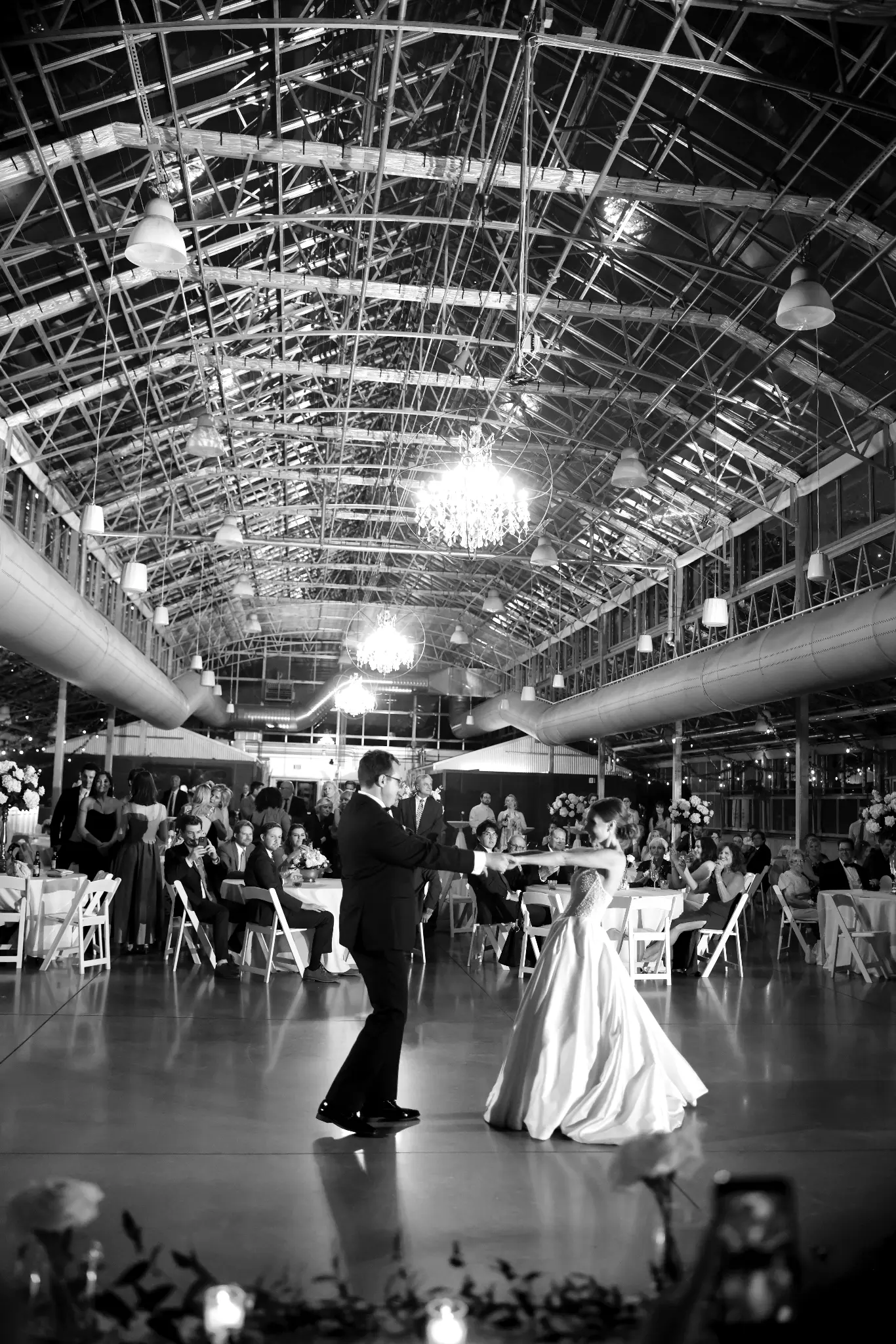 Black-and-white wide view of a wedding couple dancing in an industrial reception space