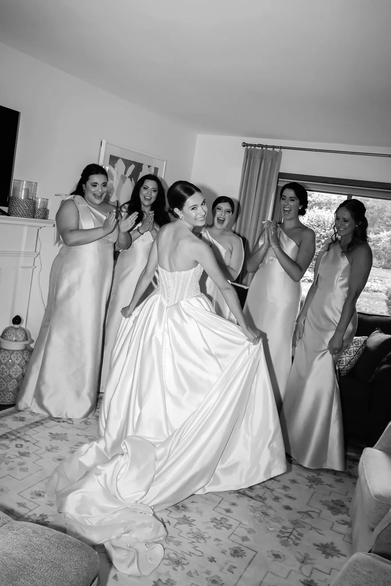 Black-and-white bride holding her gown as attendants applaud during wedding preparations
