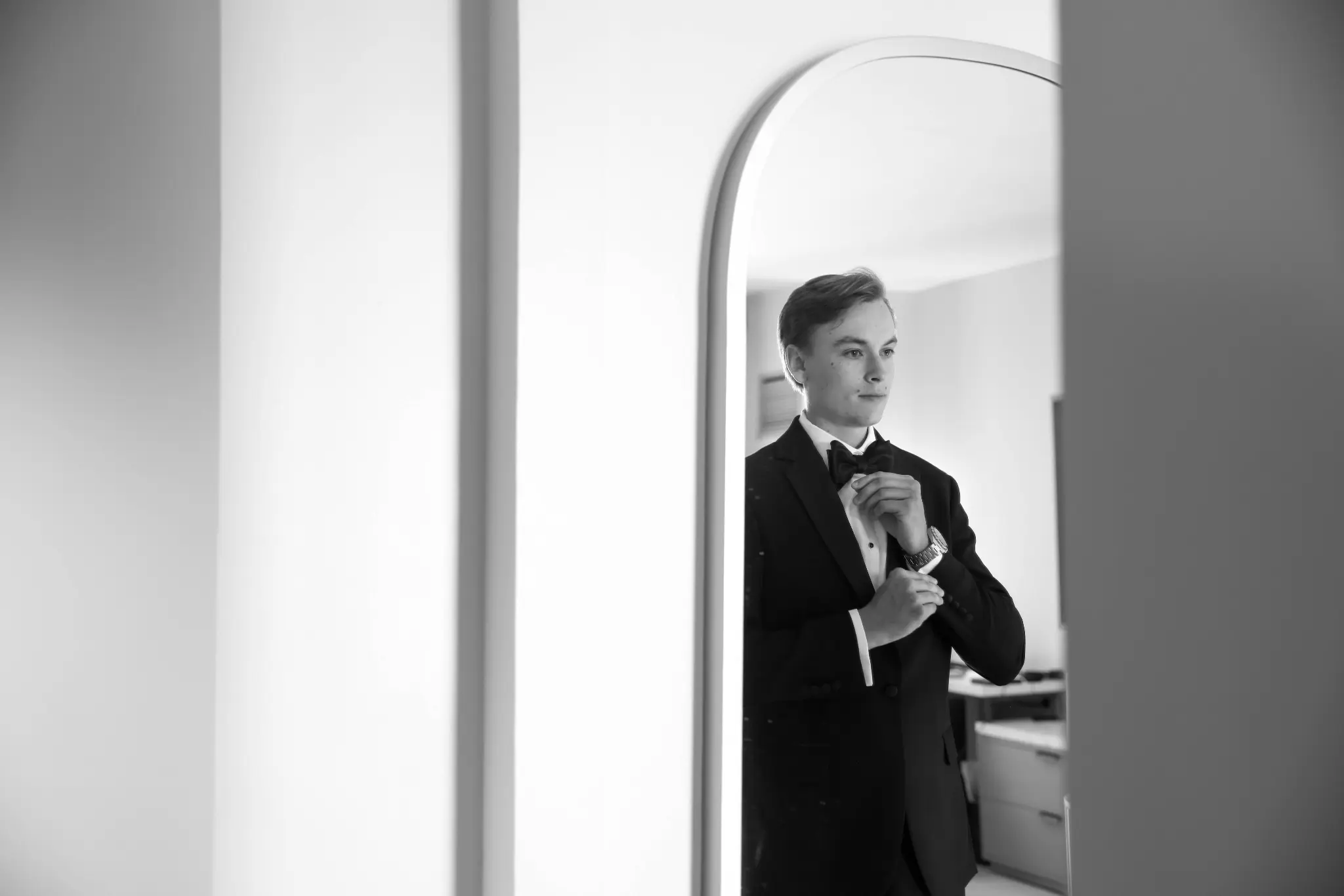 Black-and-white groom reflection adjusting his bow tie in a tall mirror before the wedding